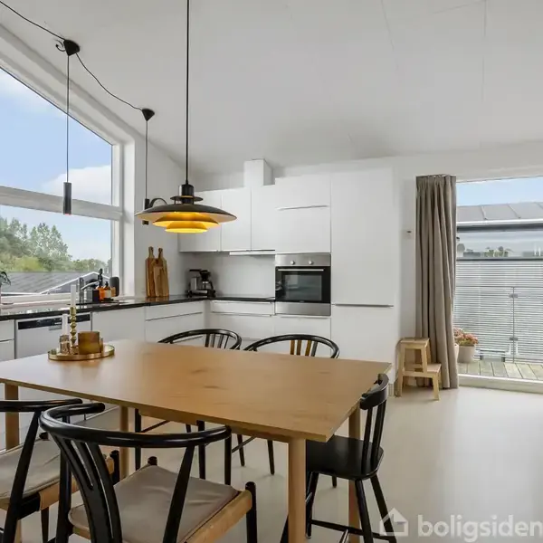 Et lyst køkken-alrum indeholder et træbord med sorte stole. En enkel gul lampe hænger over bordet. Rummet har store vinduer med udsigt til en terrasse og grønne omgivelser.