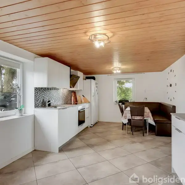 Køkken med hvidt interiør, kogeplader og ovn til venstre, lille spisebord med stole til højre, træloft og flisegulv. Store vinduer giver udsigt til træer udenfor.