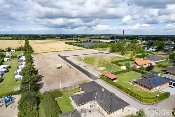 Blandet bolig- og landbrugsområde, med nybyggede huse og en stor åben mark. Området er omgivet af grønne træer, mindre veje og campingvogne. Skyer fylder den blå himmel.