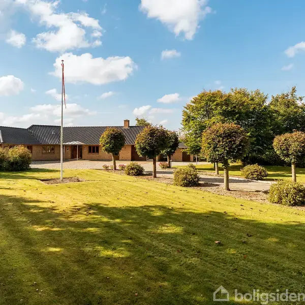 Et lavt rødt murstenshus med sort tag står stille omgivet af en velplejet have med trimmede buske. En flagstang rejser sig fra den grønne græsplæne, omgivet af blå himmel.