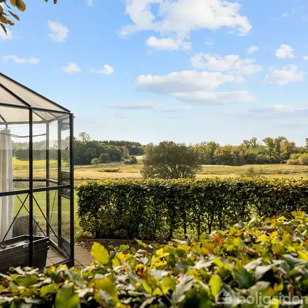 Et drivhus står stille omgivet af grønne hække i en frodig have. I baggrunden ses en vidstrakt eng med træer under en blå, skyet himmel.