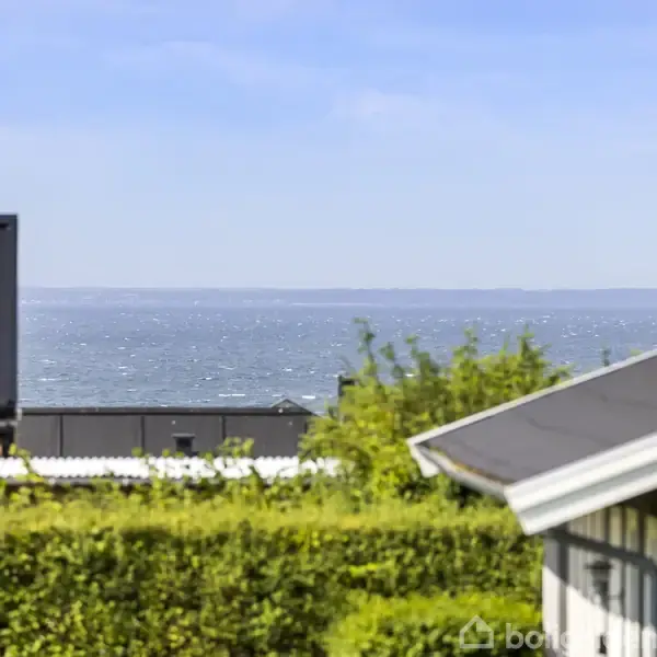 Bygninger står stille nær en hæk med udsigt over et blåt, glitrende hav under en klar himmel.