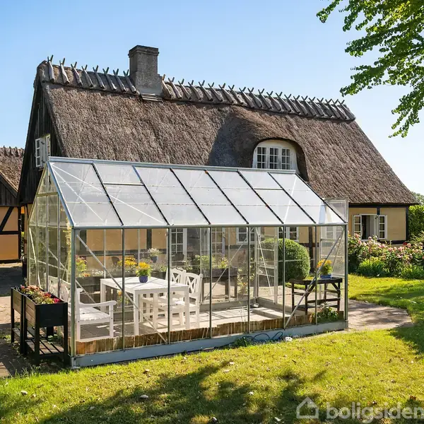 Et drivhus med glasvægge står på en terrasse ved siden af et stråtækt bindingsværkshus. I drivhuset er der hvide havemøbler. Omgivet af en frodig have med træer og buske.