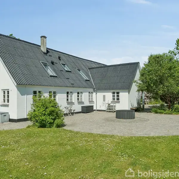 Hvidt hus med sort tag, omgivet af stenterasse og græsplæne. Havebord og stole placeret foran huset. Grønne træer og buske i baggrunden under en blå himmel.