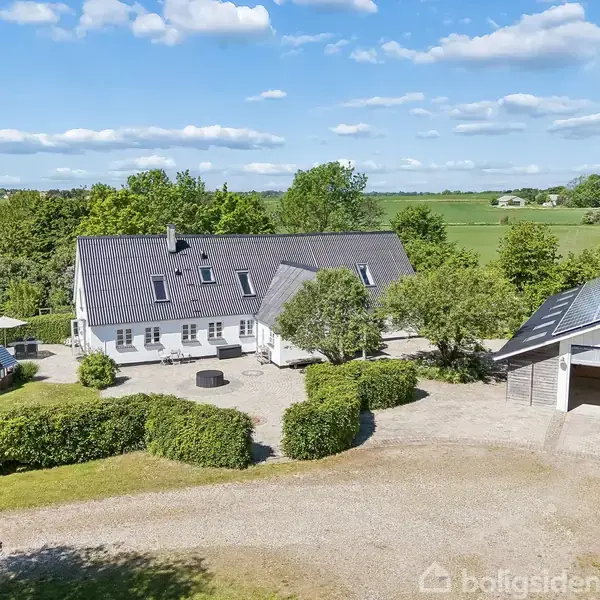 Hvidt landhus med mørkt tag står ved siden af en carport, omkranset af grusindkørsel og frodig grøn vegetation. Skyfri himmel og åbne marker skaber en rolig atmosfære.