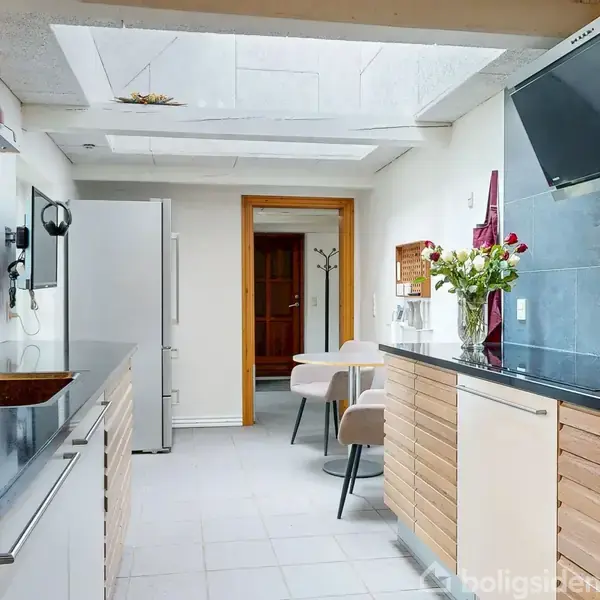 Lysegråt køkken med sorte bordplader, vask og armatur. Frisk buket blomster står på bordet. Lyse stole og et sofistikeret design under ovenlys, ved siden af hvid køleskab.