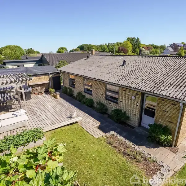 Et murstenshus i ét plan står stille i en solrig have med terrasse og pergola. Omkring er der grønne planter og udsigt over et villakvarter.