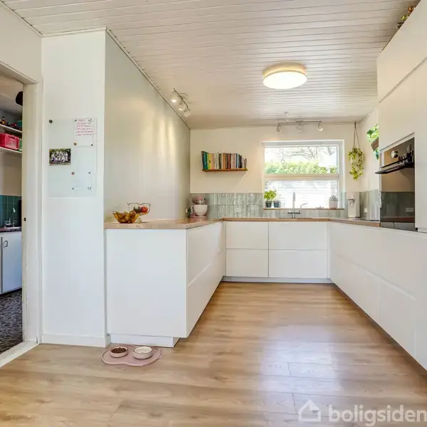 Et lyst køkken med hvide skabe og træbordplader. Et åbent gennemgangsområde til et opbevaringsrum til venstre. Vindue giver naturligt lys, og stueplanter dekorerer rummet.