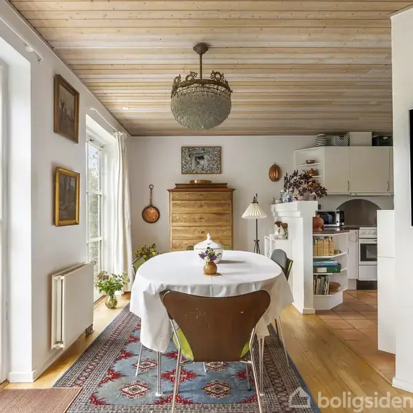 Rundt spisebord med hvid dug står i et lyst rum med trægulv og loft. Baggrunden har et skab og køkken. Store vinduer til venstre, urtepotteskjuler på bordet.