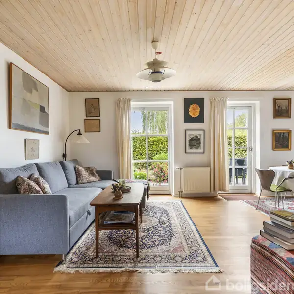 Gråt sofaarrangement står i en lys stue med træloft, vægkunst og tæppe. To glasdøre fører ud til en frodig have. Spisebord med stole ses i baggrunden.