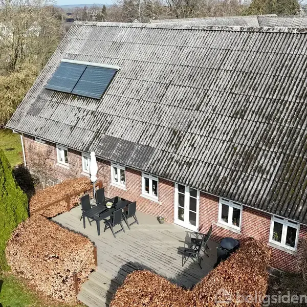Et rødt murstenshus med skrått tag og solpaneler. En træterrasse foran huset har havemøbler. Omgivet af buske og et grøn græsareal. Træer i baggrunden.