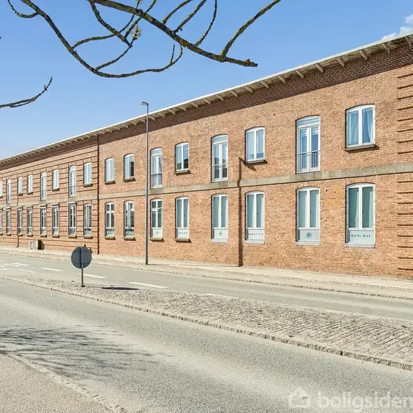 Rød murstensbygning med blå og hvide vinduer langs en rolig gade. Enkelte nøgne trægrene over vejen. Klar blå himmel uden skyer i baggrunden.