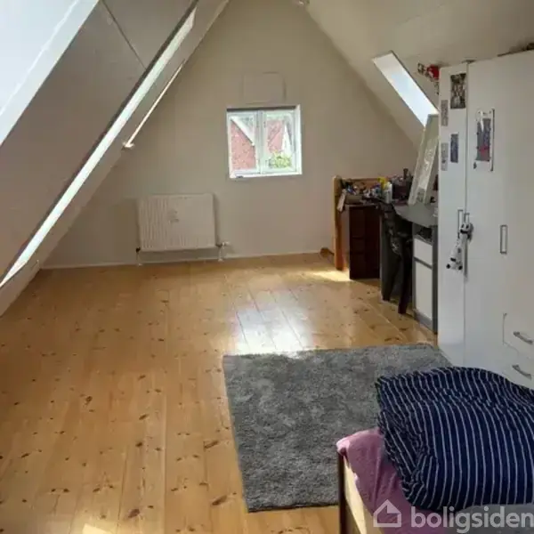 Et loftsrum med skrå vægge, trægulv og et vindue, der slipper lys ind. En radiator er monteret på væggen, og et tæppe ligger på gulvet.