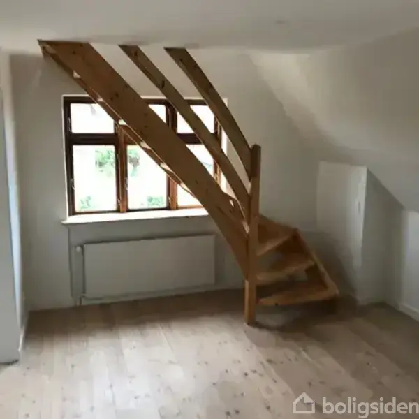 Trætrappe fører opad i en lys rum med hvidmalede vægge. En radiator er placeret under et stort vindue med udsigt til grønne omgivelser udenfor.