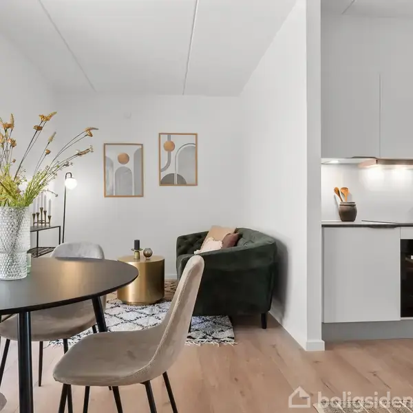 Et moderne rum viser en spisebordssæt med blomster i en vase, mens en grøn sofa står ved siden af. Rummet har en åben forbindelse til et hvidt køkken.