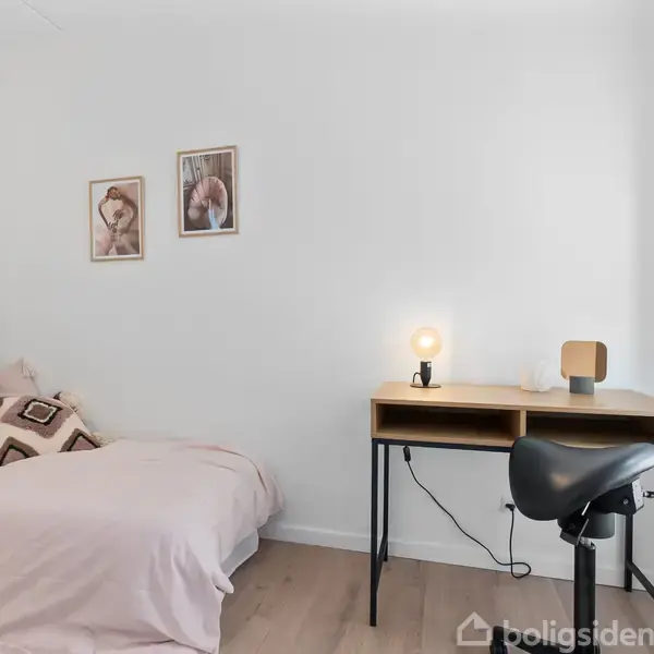 En enkeltseng med puder står ved en hvid væg, dekoreret med to billeder. Til højre en skrivepult med en lampe og stol. Trægulv og minimalistisk indretning.