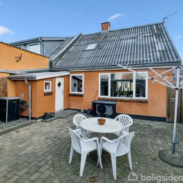 Et rundt bord med fire hvide plastikstole står på en flisebelagt terrasse foran et oranjemalet hus. En tørresnor er opstillet i hjørnet af terrassen.