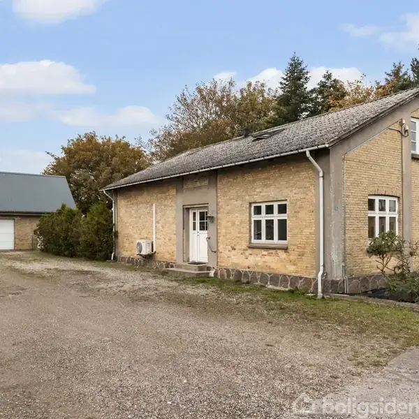 Et ældre, gulstenshus står stille ved en grusvej, omgivet af buske og træer. I baggrunden ses en garagebygning med to hvide porte under en blå himmel.