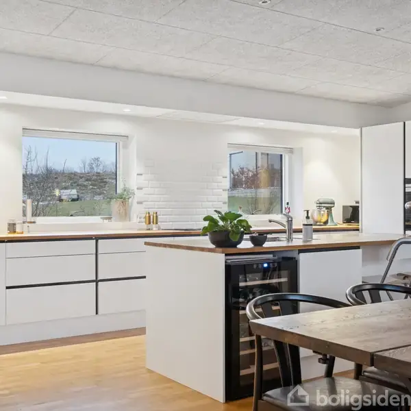 Køkken med hvidt interiør og træbordplade indeholder en køkkenø og sorte stole. Gennem de to vinduer ses træer udenfor, hvilket giver naturligt lys til rummet.