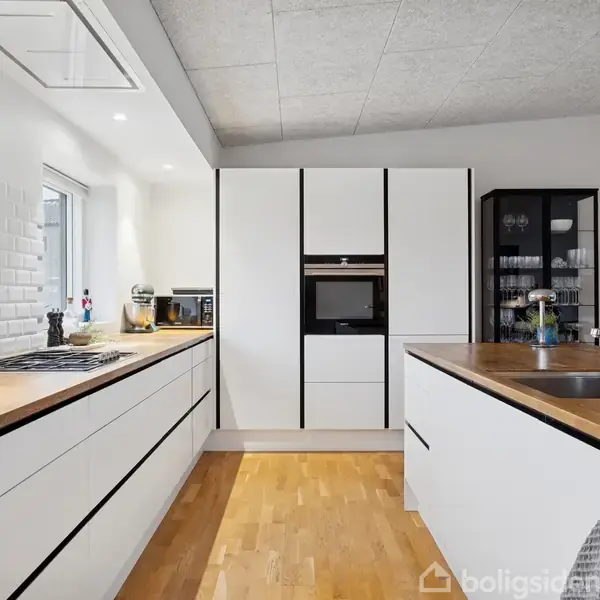 Hvidt, moderne køkken med træbordplader, indeholder indbyggede apparater og kogeplade. Skyllevask er placeret på en køkkenø. Rummet har lyst trægulv og hvide flisevægge.