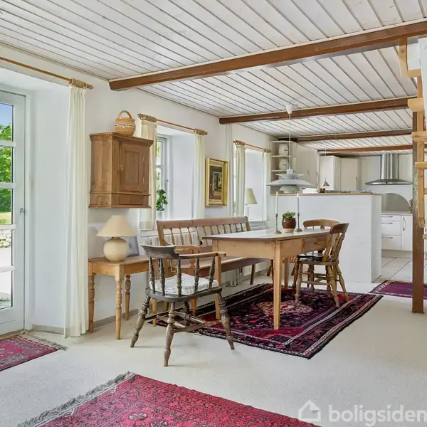 Spisebord med stole står i en lys stue med røde tæpper. Rummet har hvide vægge, træloft, og store vinduer med hvide gardiner. Trappe fører op til en anden etage.