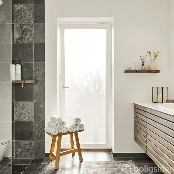 Badeværelse med brusekabine er til venstre, med mørke fliser og shampoo samt bruser. En skammel med sammenrullede håndklæder står i midten. Til højre er der en vask med spejl, hylder og lysdekorationer.