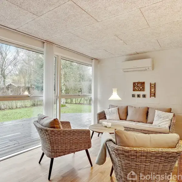 Stue med flettede møbler, hvor sofa og stole vender mod et sofabord; store vinduer viser en terrasse og have udenfor, mens lamper lyser indendørs.