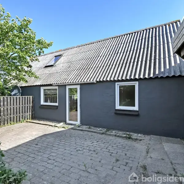 Et gråt hus med skråt tag står i et brolagt gårdhaveområde. To vinduer og en dør vender mod gårdhaven. Til venstre ses et sort plankeværk og et træ.