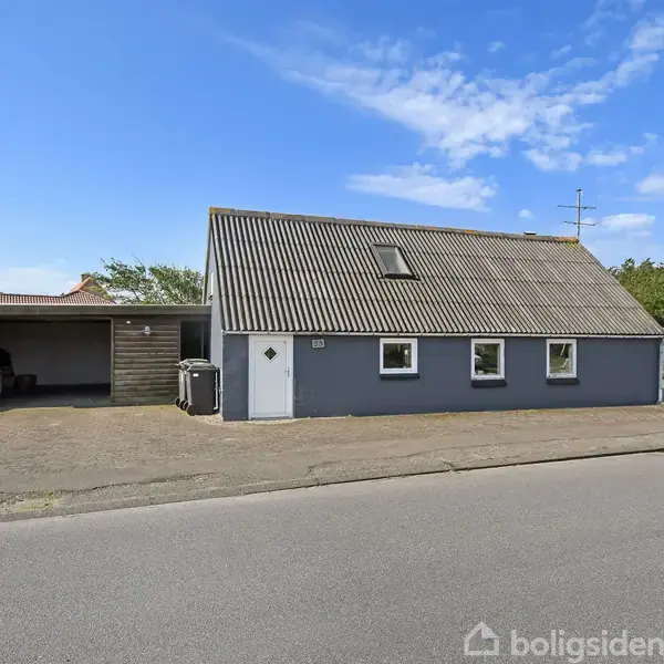 Et gråblåt hus med skråt tag står stille ved en vejkant. Der er fire vinduer og en dør foran. Til venstre, en carport med åbent rum.