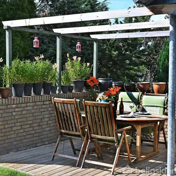 Terrassemøbler står omkring et dækket træbord med vinflasker og glas. Bagved ses blomsterpotter på en murstensvæg under en pergola med lamper. Omgivet af grønne buske og træer.