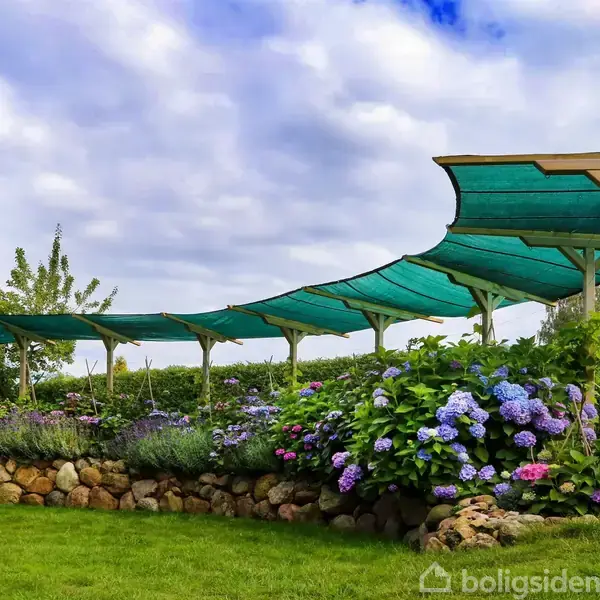 Grønne skygge-sejl strækker sig over en have med blomsterbede og stengærder, omgivet af tætte buske og træer under en blå, let overskyet himmel.