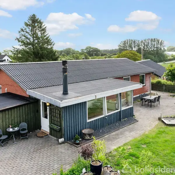 En moderne enfamilieshus med rødt og sort tag, terrassen har to siddeområder. Omgivet af en grøn have og marker, under en blå, skyet himmel.