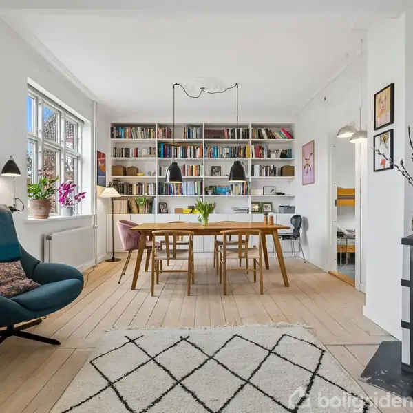 Stue med pejs; moderne stol ved trebord og tæppe, omgivet af bogreoler, planter og kunst på væggen, skaber en hyggelig og lys atmosfære med lamper og trægulv.