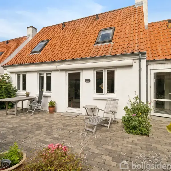 Hvidt hus med orange tegltag står i en gårdhave. Havemøbler står på stenbelægning omgivet af grønt løv. Vinduer har hvide rammer, og der er en glaspaneldør.