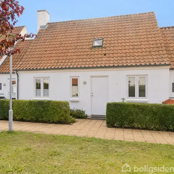 Hvidt rækkehus med rødt tegltag står stille i sollys. Foran er en velholdt hæk og en flisebelagt sti. Græsplæne og buske udgør omgivelserne.