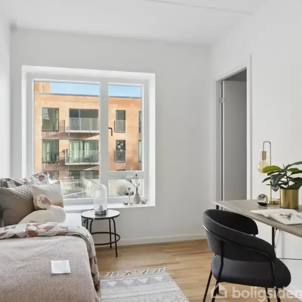 Værelse med seng og skrivebord. Store vinduer giver udsigt til moderne lejlighedskompleks udenfor. Indretning inkluderer puder, en plante, og en lampe på et lyst trægulv.