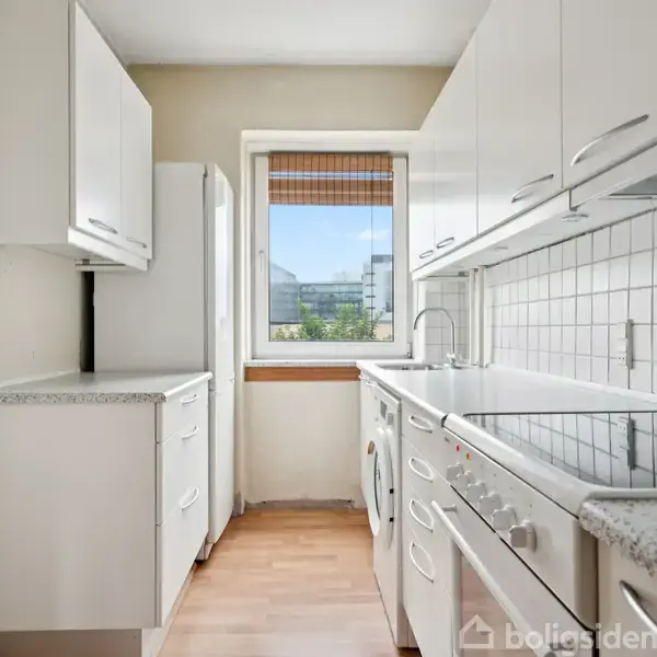 Køkken med hvide skabe og lys træbordplade, indeholder komfur og vaskemaskine. Sollys strømmer ind gennem vinduet, der åbner ud til en udsigt over bygninger.