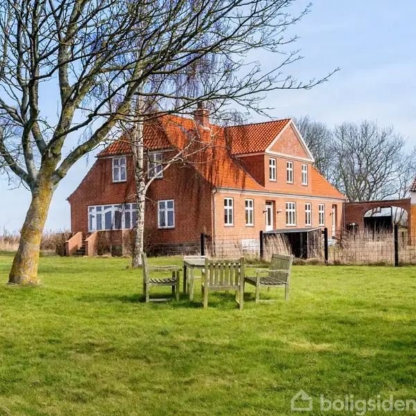Et rødt murstenshus med en rød tag sidder blandt grønne græsplæner, med træer i nærheden. Et sæt havemøbler står på plænen foran huset, omgivet af åben natur.
