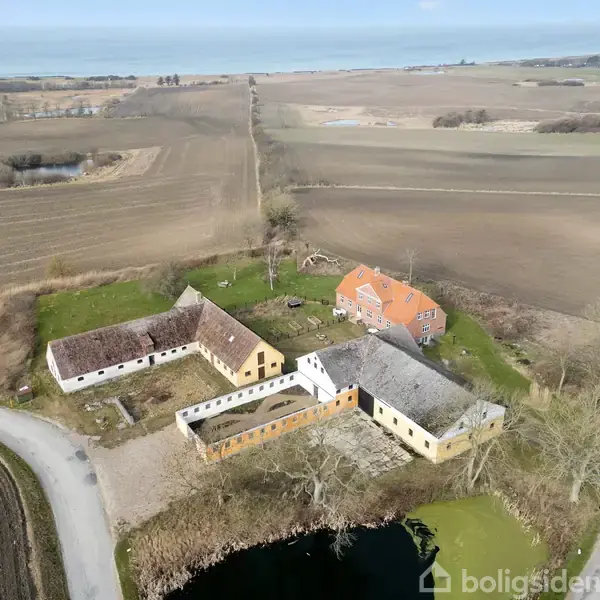 Gårdkompleks med rødt og hvidt murstenshus ligger ved en sø, omgivet af opdyrkede marker og snoede veje under en klar himmel.