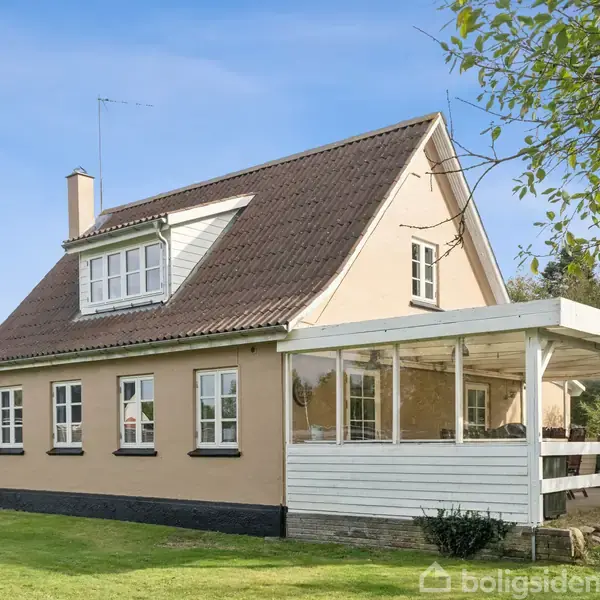 En beige villa med mørkt skråt tag, store vinduer og en overdækket veranda, står på en velholdt græsplæne, omgivet af træer under en klar blå himmel.