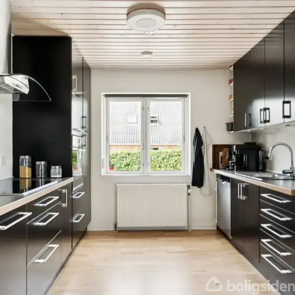Et moderne køkken med sorte skabe og stålgreb på begge sider af rummet. En stor stål-køleskab står til højre, og i midten er et vindue med udsigt til en have.