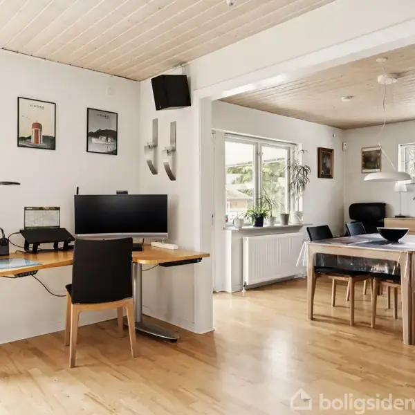 Et skrivebord med en computer og printer står ved en væg dekoreret med billeder. I baggrunden ses et spisebord med stole i et lyst rum med vinduer og træloft.