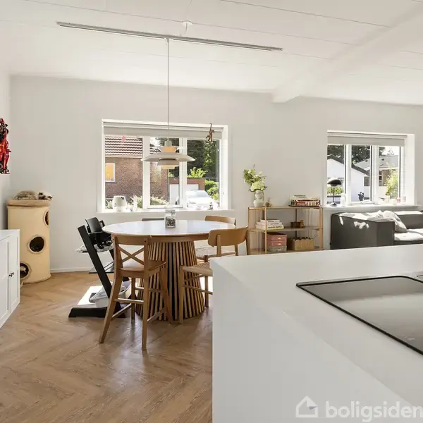 Et lyst køkken-alrum med spisebord og stole, placeret ved siden af vinduer der giver udsigt til en have. Køkkenet har en hvid indretning og en sort kogeplade.