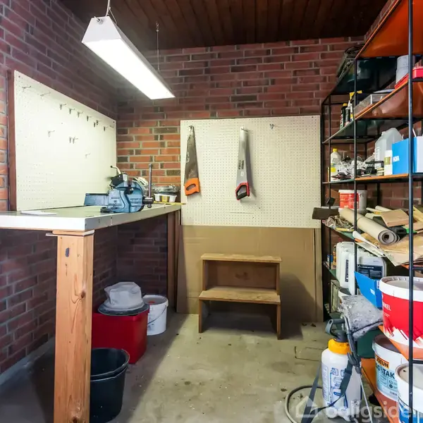 Et velorganiseret værksted med arbejdsbord og værktøjstavle. Bakker står op ad væggen. Hylder til højre opbevarer maling og værktøj. Væggene er af røde mursten.