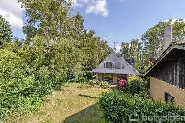 Et A-formet hus står i en frodig have med store træer og buske. Det er en solrig dag med spredte hvide skyer på himlen.