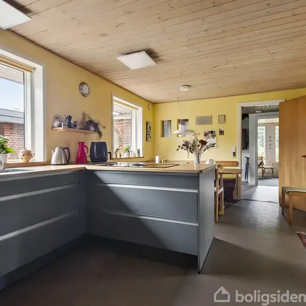 Køkken med grå skabe i aktivitet med køkkenudstyr og planter på bordpladen. I baggrunden ses et spiseområde med træmøbler, omgivet af lyse vægge og store vinduer.