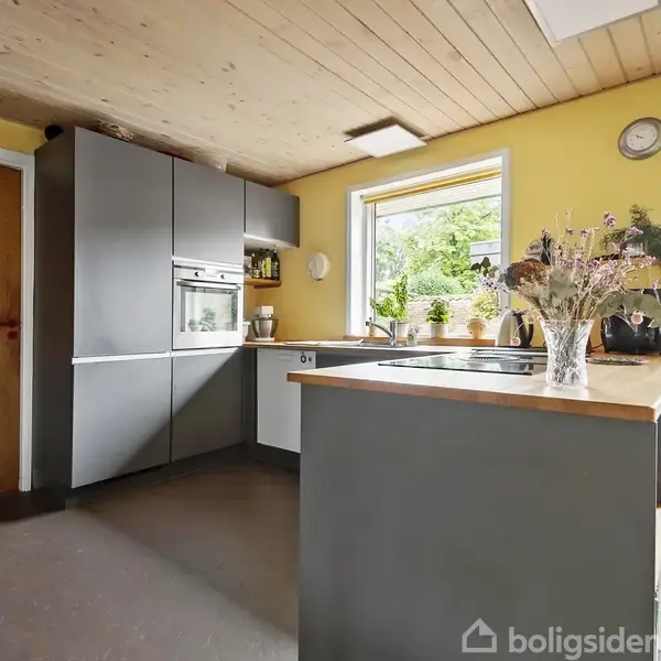 Køkkenbord med vask og blomsteropsats står i centrum. Vinduet viser udsigt til træer. Grå skabsvæg med indbygget ovn. Gulmalede vægge skaber en varm atmosfære.