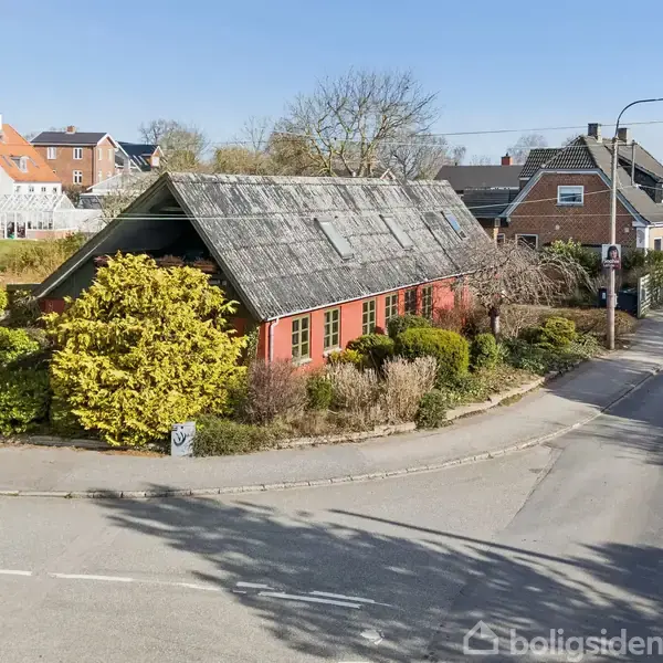 Et gammelt træskur med skrånende tag er omgivet af grønne buske og placeret ved en vejskrids i et beboelsesområde. Baggrunden viser flere huse i en bymæssig kontekst.