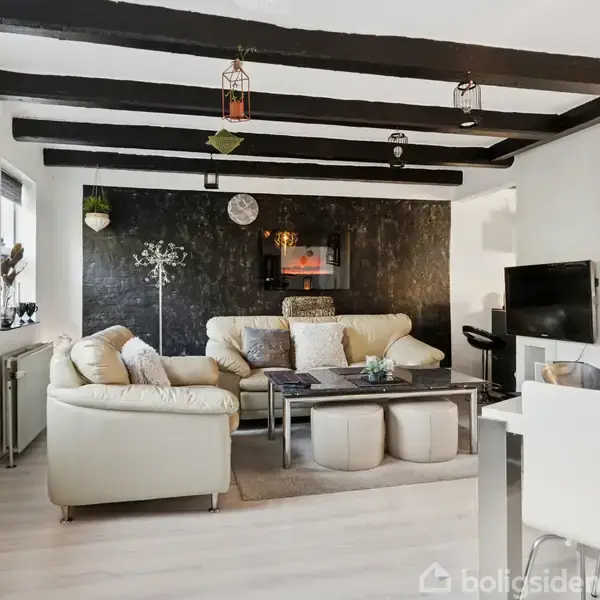 Sofa i lys farve står i en stue med sortmalet væg og træbjælker i loftet. Der er et sofabord med puder under, og et TV hænger på væggen.