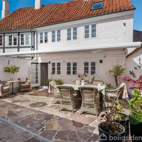 To gårdhavemøbler står på en stenbelagt terrasse foran et hvidt hus med rødt tegltag. Omgivet af planter og blomsterdesser, skaber det en hyggelig, indbydende atmosfære.